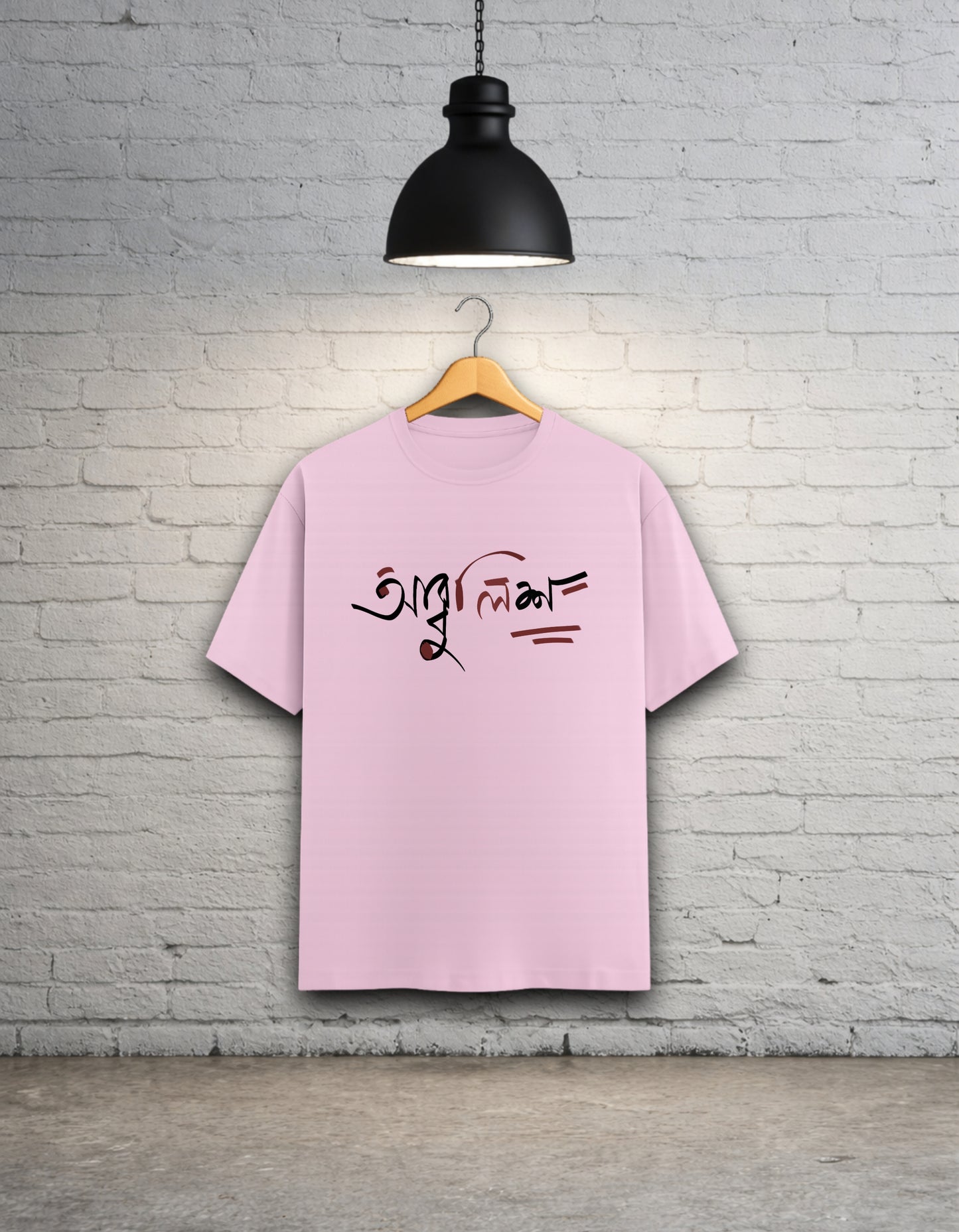 Baby pink t-shirt with text design 'Abbuleesh' on a wooden hanger against a white brick wall.