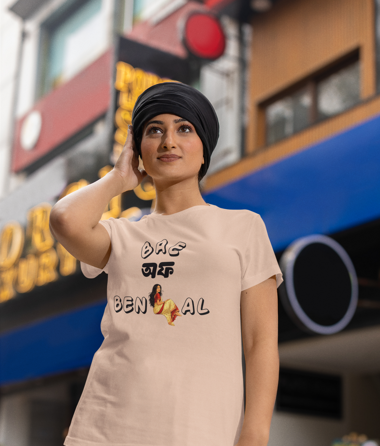 Woman wearing a peach t-shirt with text "Bae of Bengal' graphics, standing in front of a building with signs.