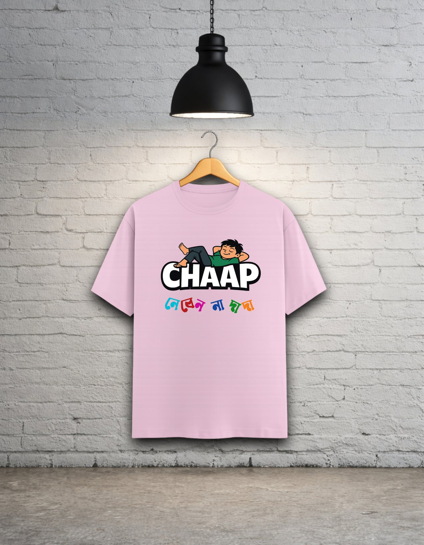 Baby pink t-shirt with 'CHAAP nebenna dada' bengali graphic design hanging on a brick wall.